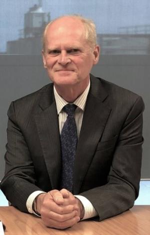 Dr Russell G Smith sits on a desk facing the camera with his hands clasped together and a resolute expression on his face